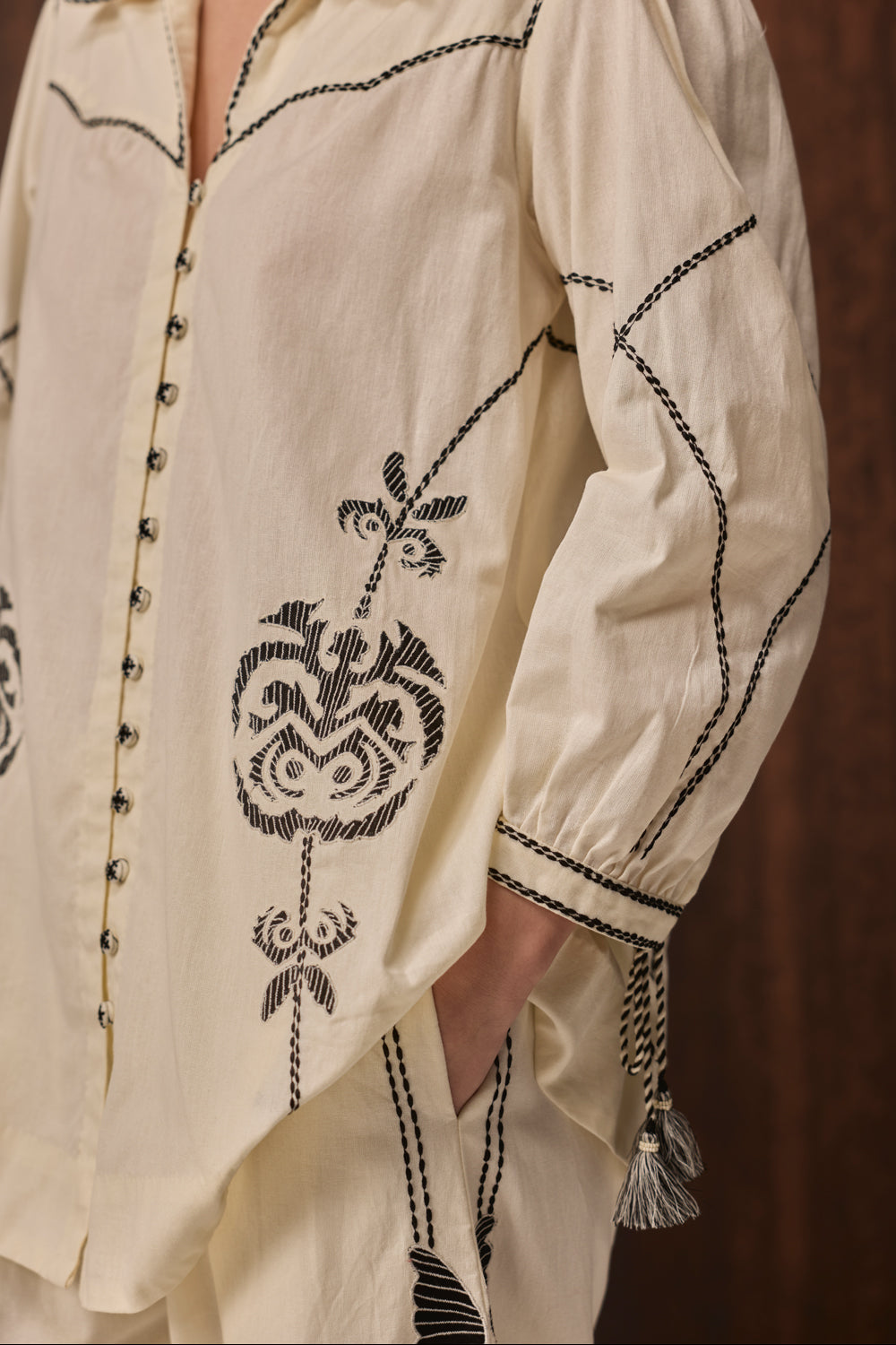 Close-up of a cream-colored embroidered shirt with black stitched patterns, decorative buttons, and tassel ties on the cuffs, worn by a model.