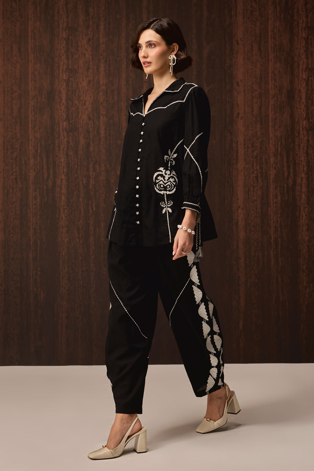 A woman models a black two-piece embroidered outfit consisting of a long-sleeve button-front top and matching cropped pants, with white contrast stitching and ornate embroidery details. She is posed against a dark wood-paneled background.