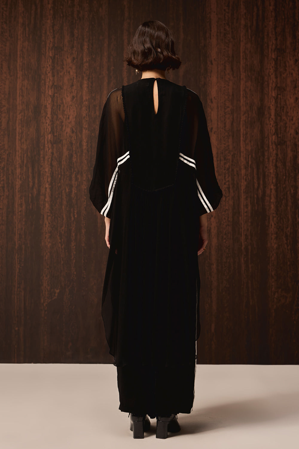 A woman viewed from behind wearing a long black dress with sheer three-quarter sleeves and white stripe accents on the cuffs, standing against a dark wood-paneled background.