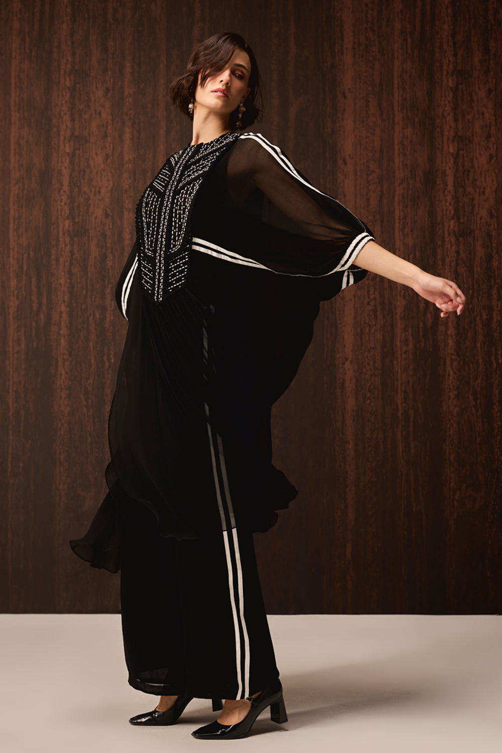 A woman modeling a black flowing maxi dress with white stripe details on the sleeves and legs, and silver geometric embroidery on the bodice, set against a dark wood-paneled background.