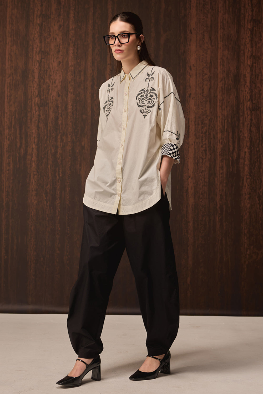 A woman modeling a cream-colored button-up shirt with black floral embroidery on the chest and sleeves, paired with black wide-leg trousers and black pointed-toe heels, set against a dark wood-paneled background.
