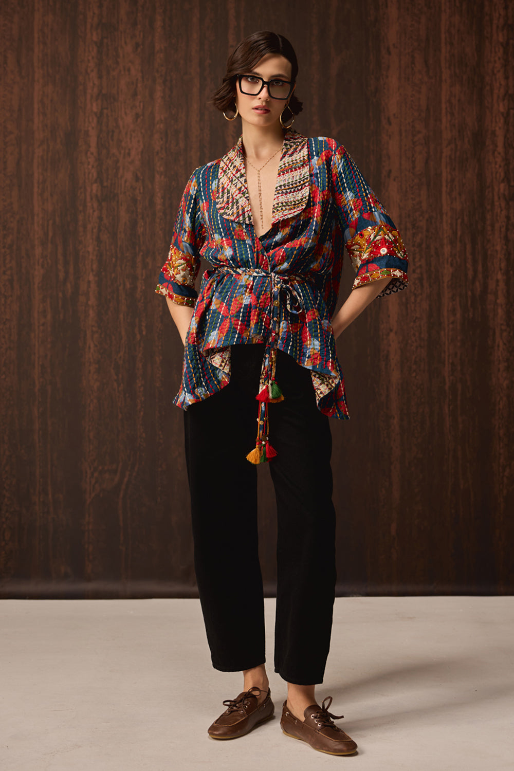 A model wearing a colorful patterned wrap-style shirt with tassel ties, black pants, and brown boat shoes, standing against a dark wood-paneled background.