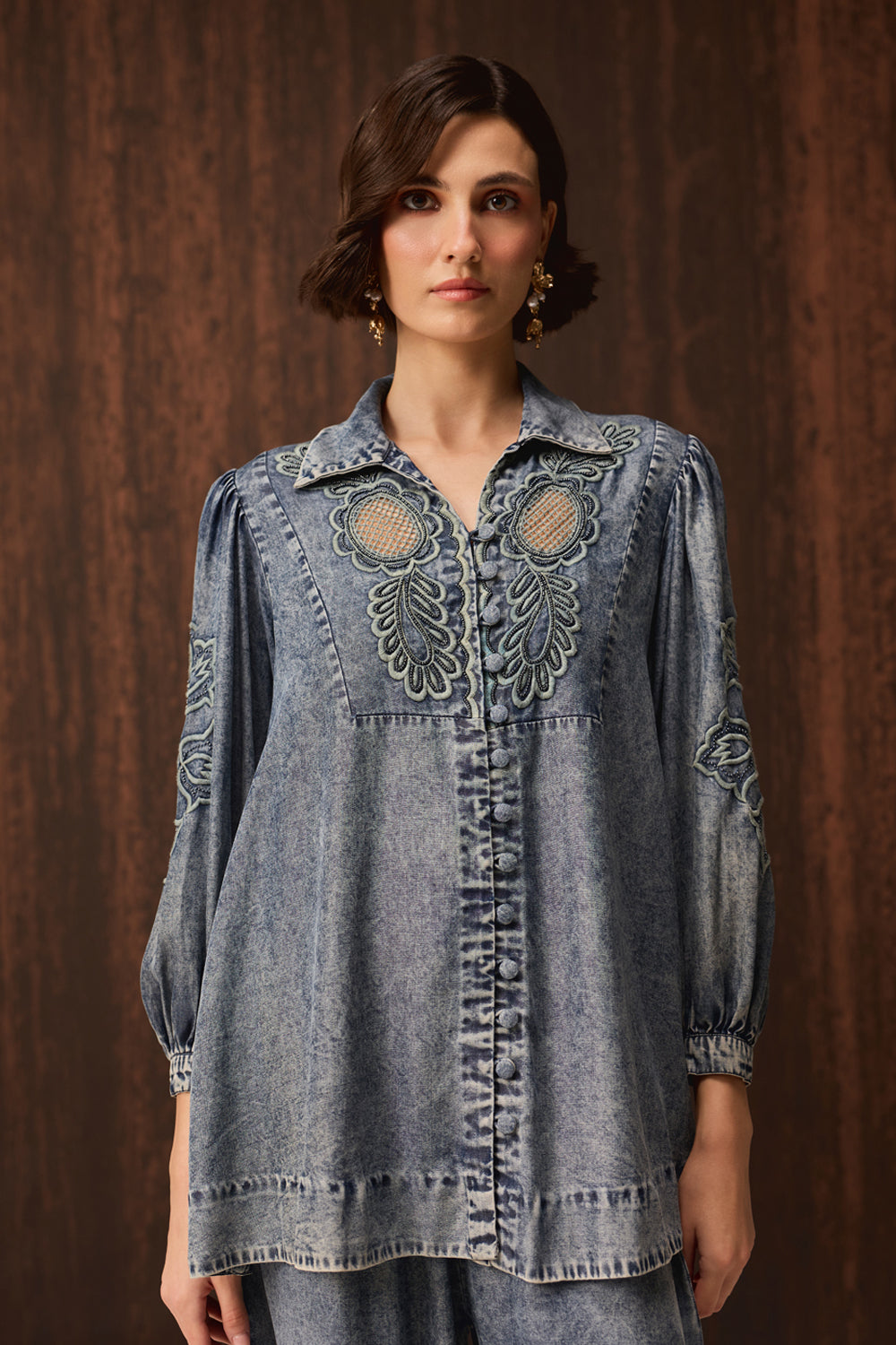 A woman wearing a blue denim long-sleeve button-up shirt with intricate embroidered detailing on the chest and sleeves, posed against a dark brown wood-paneled background.