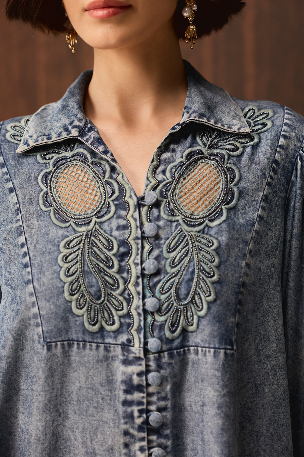 A close-up of a model wearing a blue denim button-up shirt with intricate embroidered floral details on the chest.