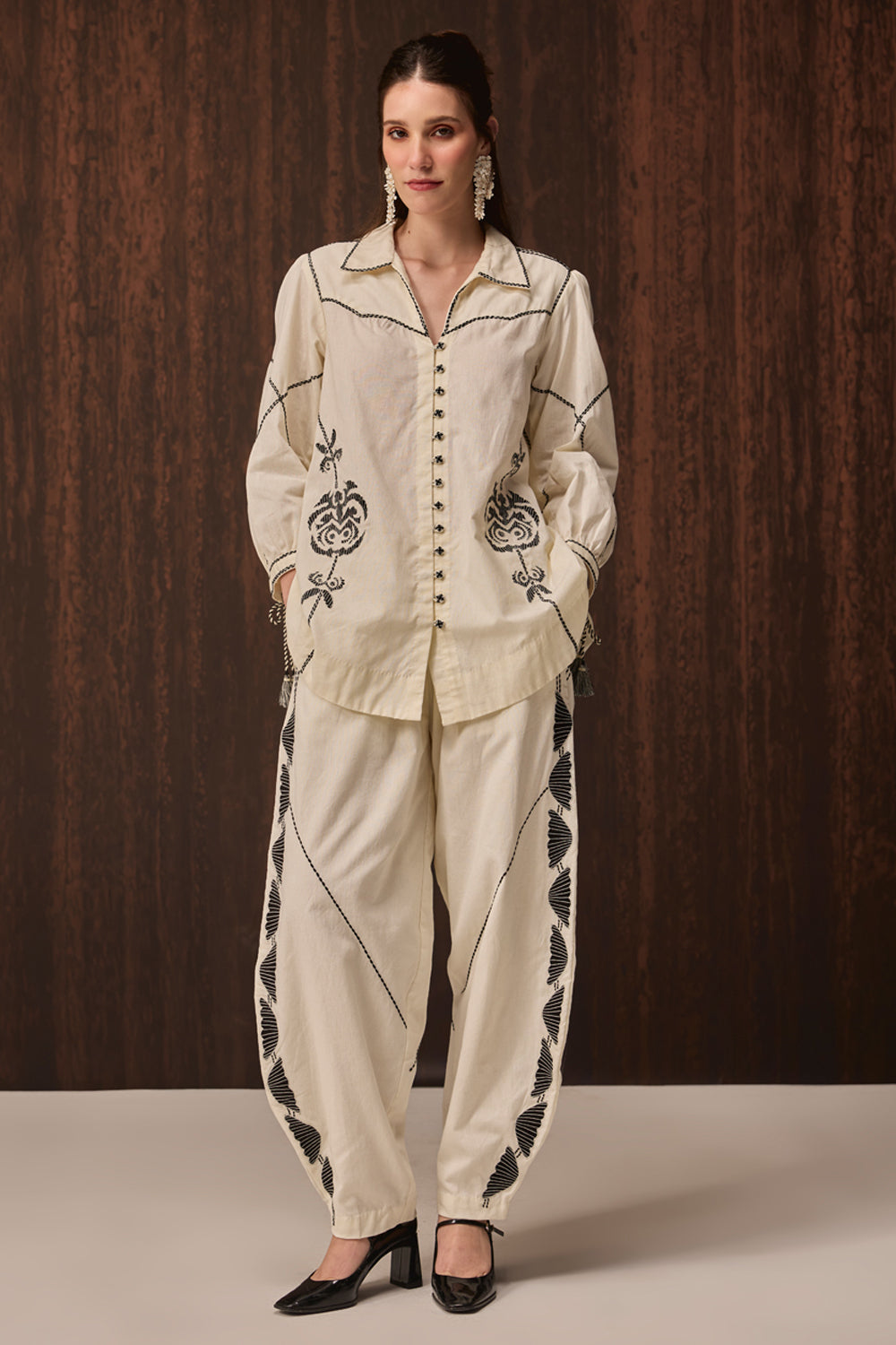 A woman models a cream-colored two-piece western-style set consisting of a long-sleeve button-up shirt and matching pants, both decorated with black floral embroidery and geometric patterns, set against a dark wood-paneled background.