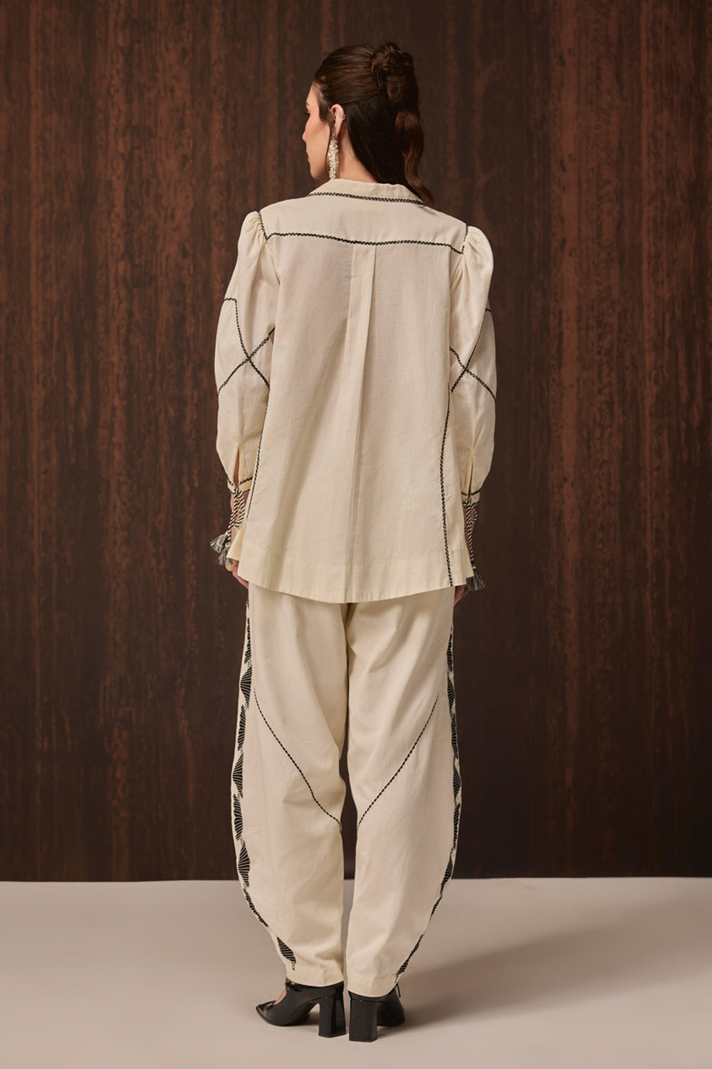 A model viewed from behind wearing a cream-colored two-piece suit with black contrast stitching and fringe details, set against a dark wood-paneled background.