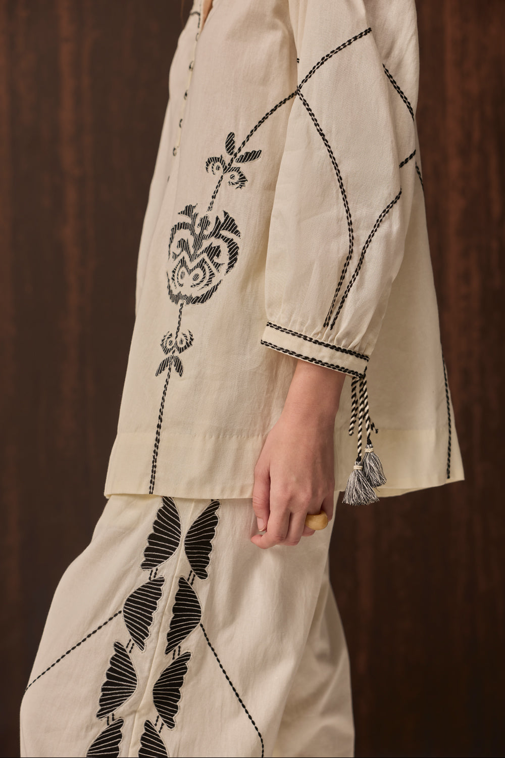 A close-up of a person wearing a cream-colored embroidered kurta and matching trousers with black floral embroidery and black stitched details, set against a dark brown wooden background.
