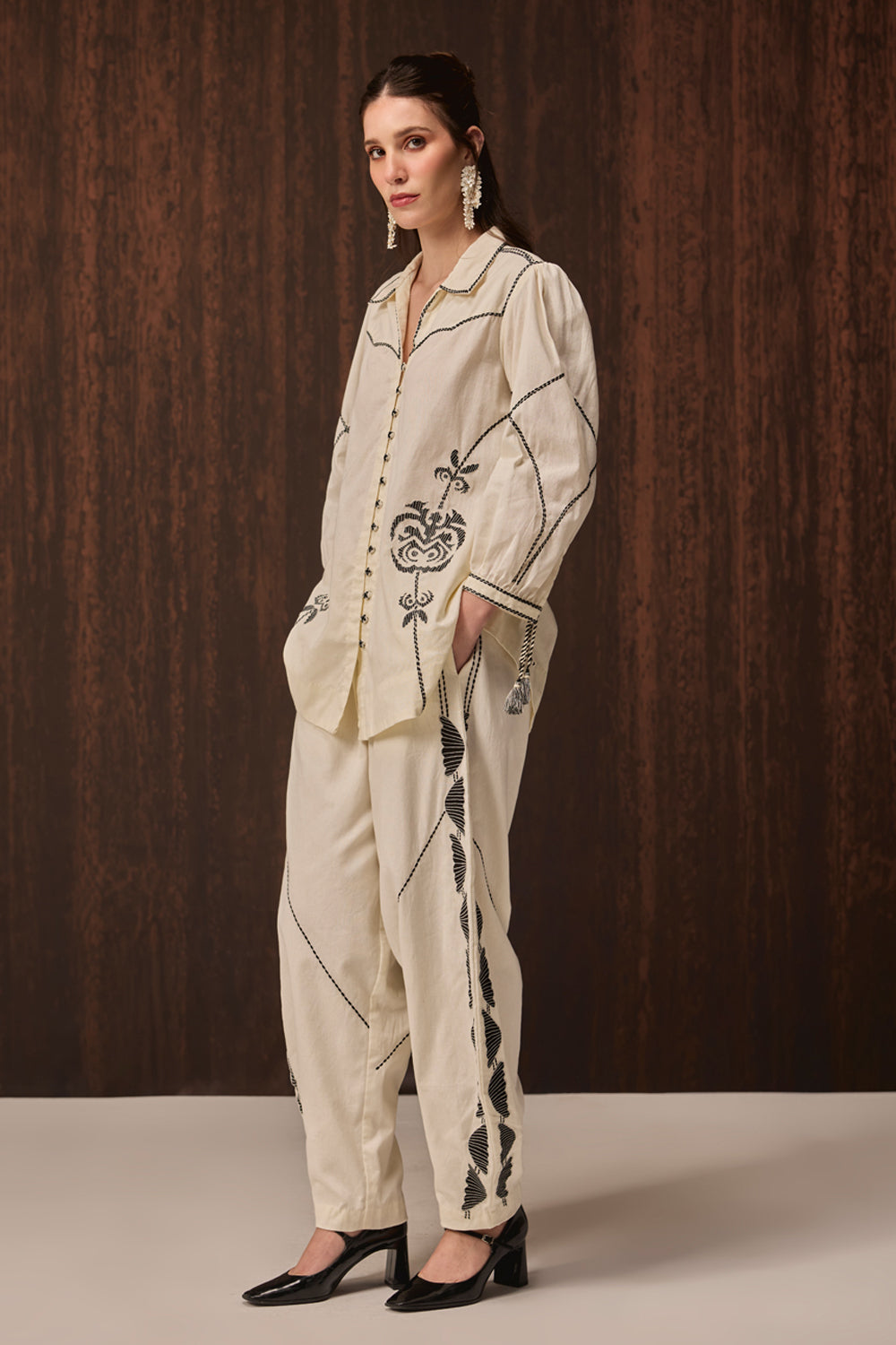 A woman models a cream-colored two-piece pajama set with black embroidered details, standing against a dark wood-paneled background.