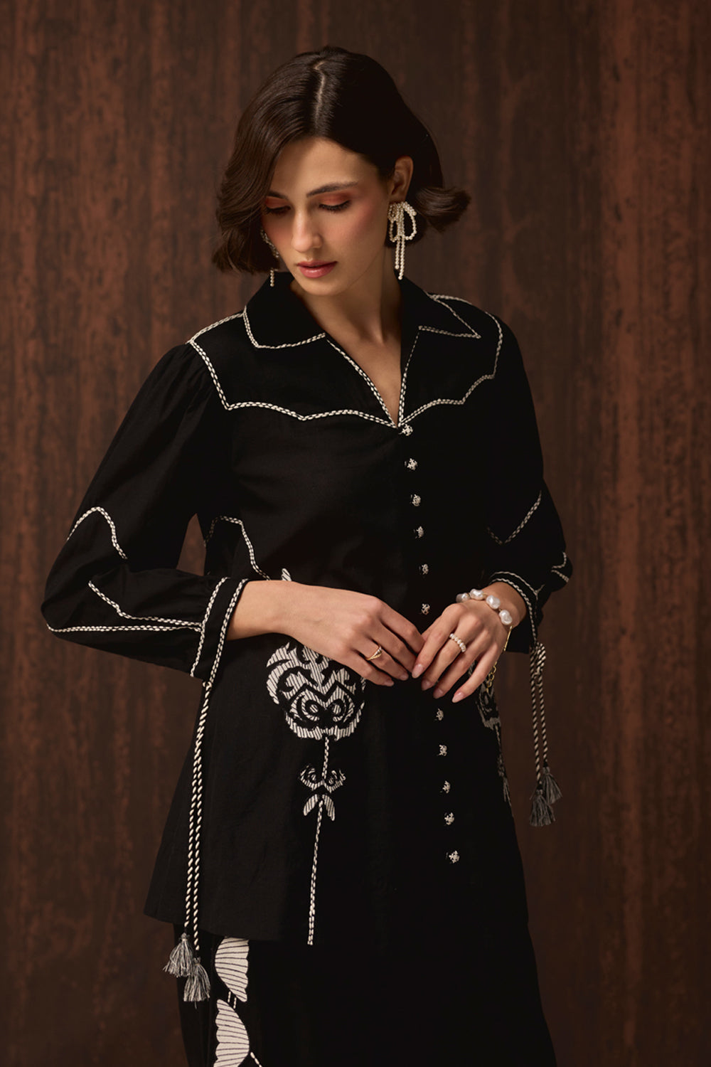 A woman wearing a black long-sleeve embroidered tunic top with silver-toned embroidery and tassel details, posed against a dark brown wood-paneled background.