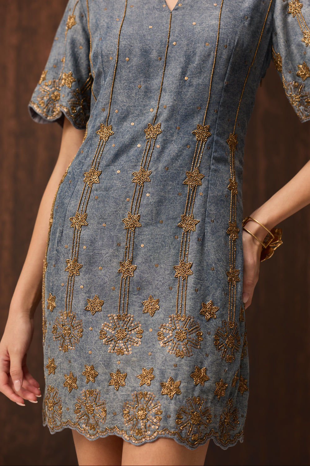 A close-up of a woman wearing a short-sleeve blue-gray dress with intricate gold embroidery and beadwork, set against a dark brown background.
