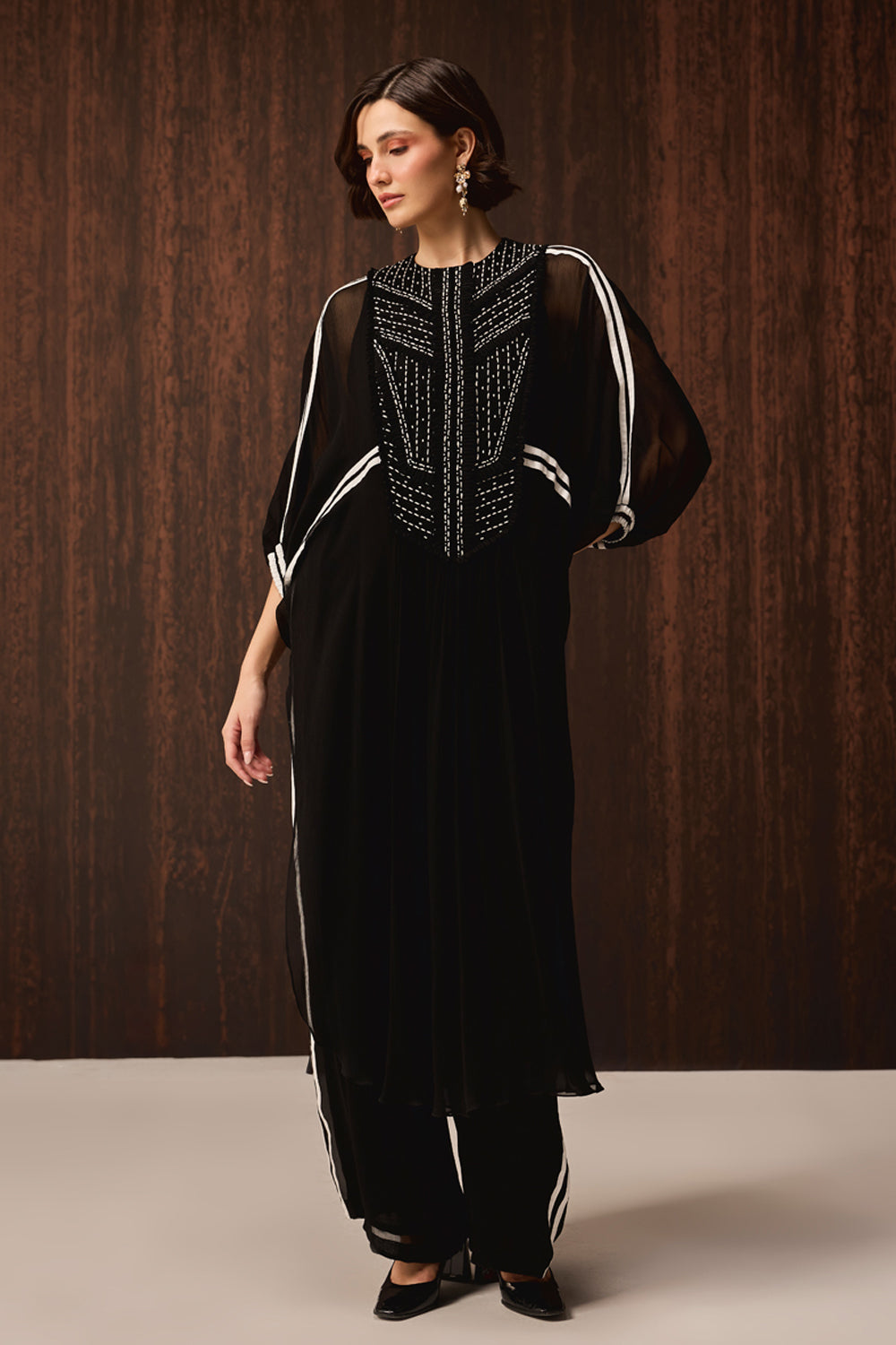 A woman models a black sheer tunic with white stripe details and a heavily embellished embroidered neckline, paired with matching black wide-leg trousers with white stripes, set against a dark wood-paneled background.