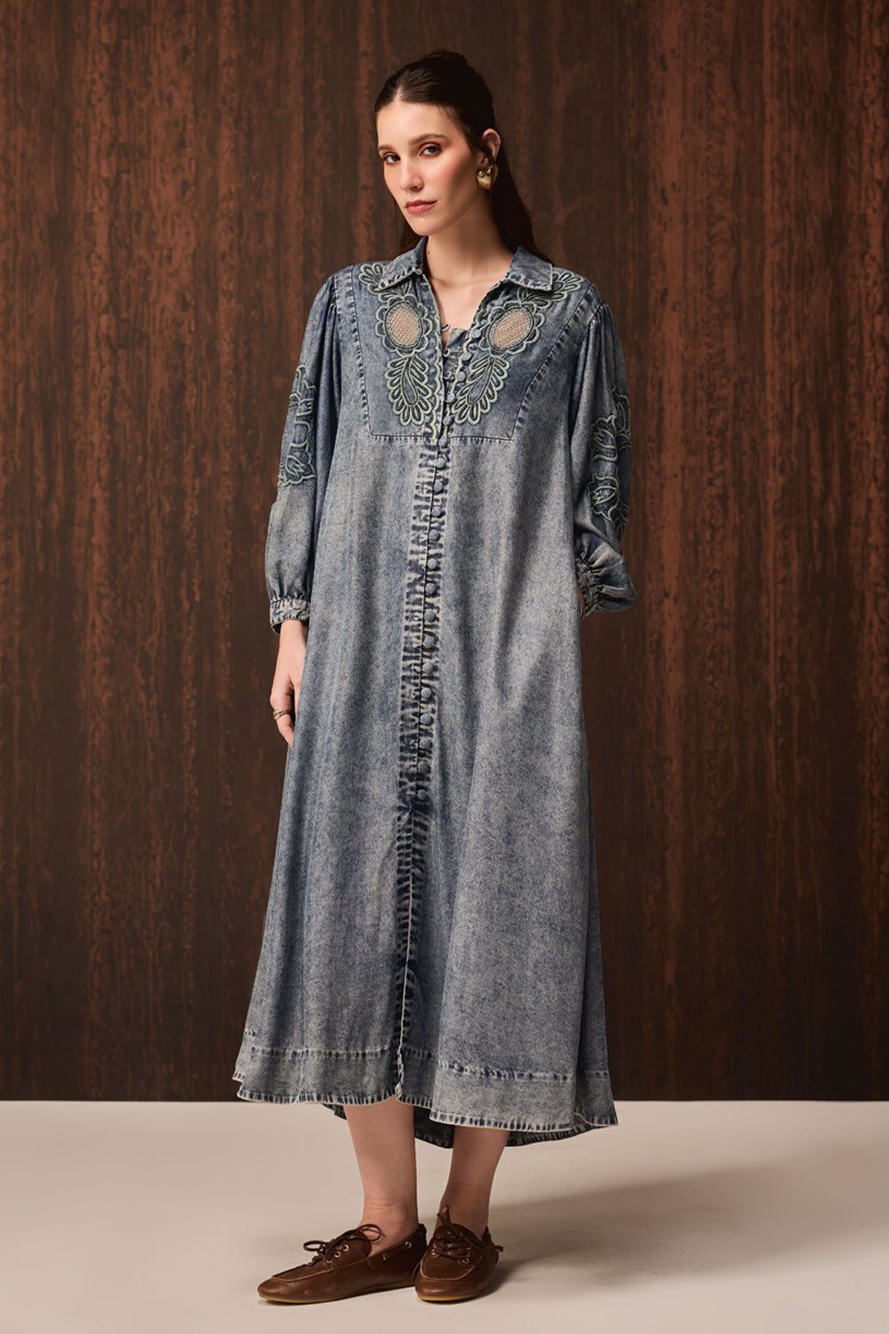 A woman modeling a long-sleeved, button-front denim dress with decorative embroidery and ruffle details, paired with brown leather loafers, set against a dark wood-paneled background.