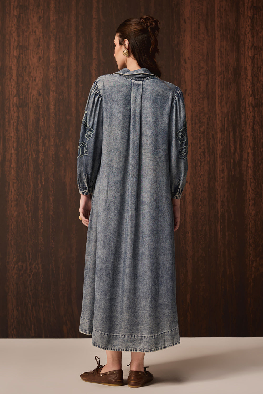 A woman viewed from behind wearing a long, wide-sleeved denim tunic dress with gathered sleeves and a raw hem, paired with brown leather shoes, set against a dark wood-paneled background.