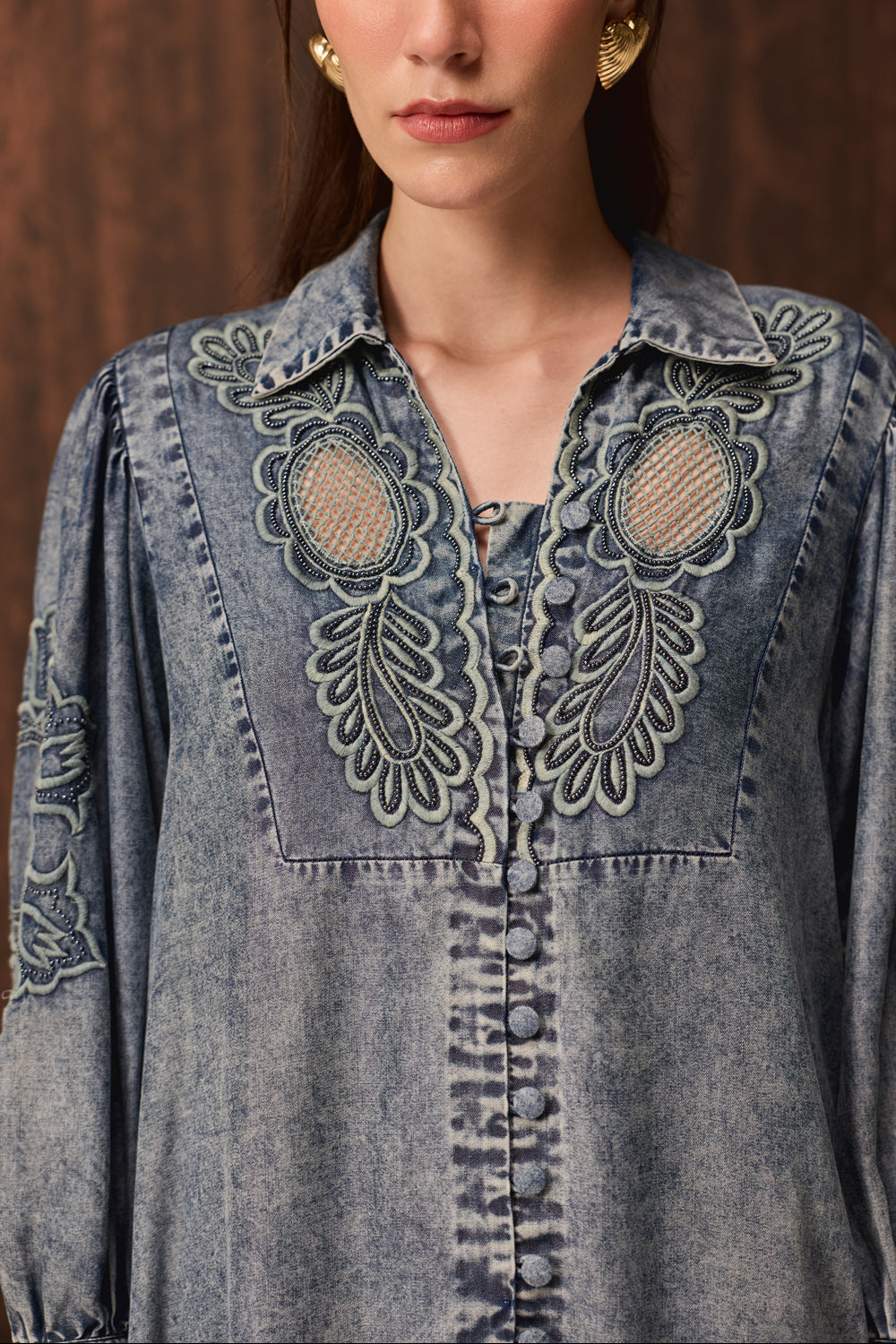 A close-up of a model wearing a blue acid-wash denim shirt jacket with intricate floral embroidery and mesh detailing on the chest, set against a dark brown textured background.