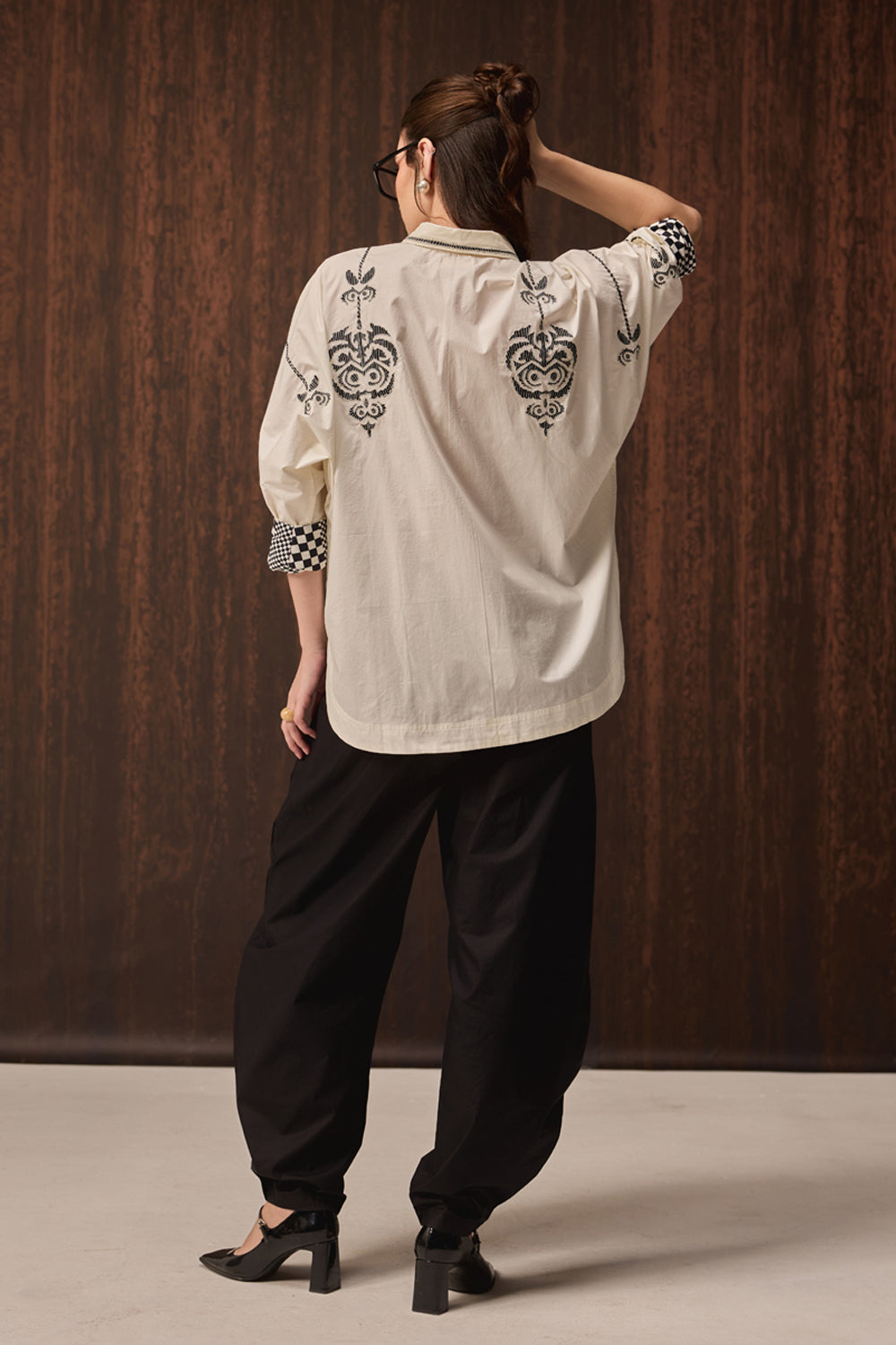 A woman shown from the back wearing a cream-colored embroidered shirt with black paisley patterns, paired with black baggy pants and black heels, standing against a dark wood-paneled wall.
