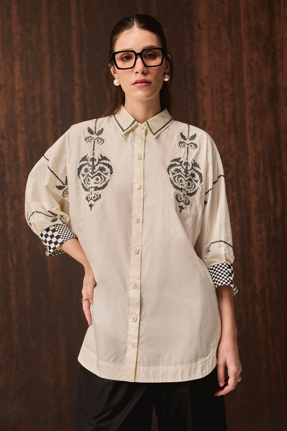 A woman wearing a cream-colored button-up shirt with black embroidered patterns on the chest and black-and-white checkered cuffs, posed against a dark wood-paneled background.