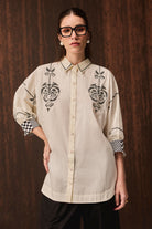 A woman wearing a cream-colored button-up shirt with black embroidered patterns on the chest and black-and-white checkered cuffs, posed against a dark wood-paneled background.