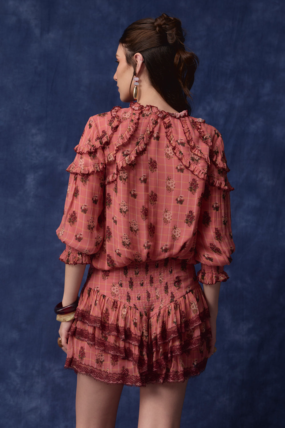 A model shown from the back wearing a dusty rose pink two-piece set consisting of a tiered ruffle blouse and matching mini skirt, both with a small floral pattern. The background is a textured dark blue studio backdrop.