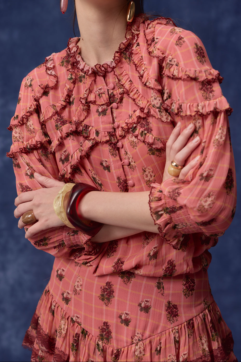 A close-up of a model wearing a pink floral long-sleeve blouse with multiple ruffle details, accessorized with gold and brown statement rings and bangles, set against a dark blue textured background.