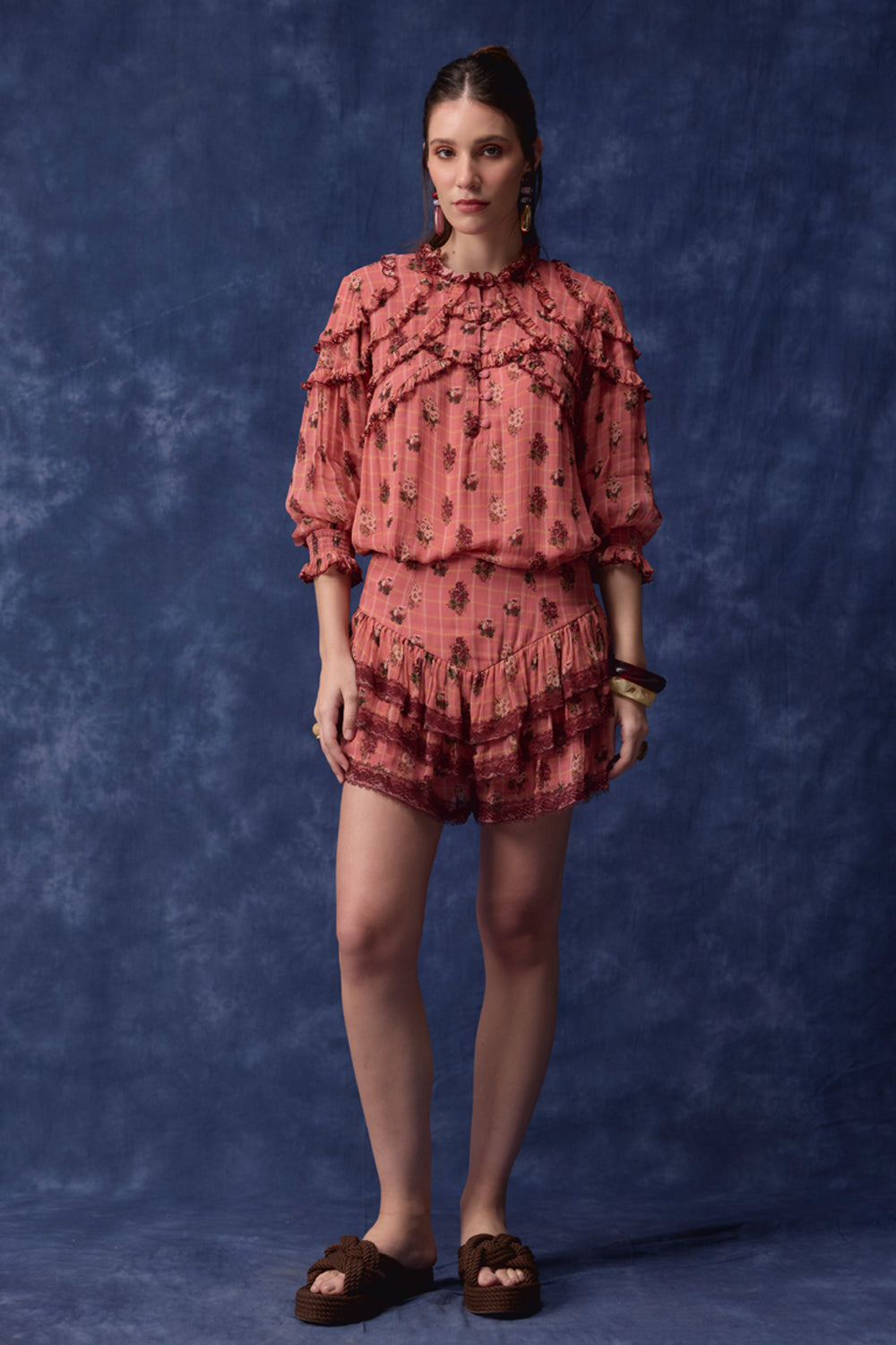 A model stands against a mottled blue studio backdrop wearing a matching two-piece outfit consisting of a ruffled long-sleeve top and shorts in a dusty rose pink color with a small floral print, paired with brown slide sandals.