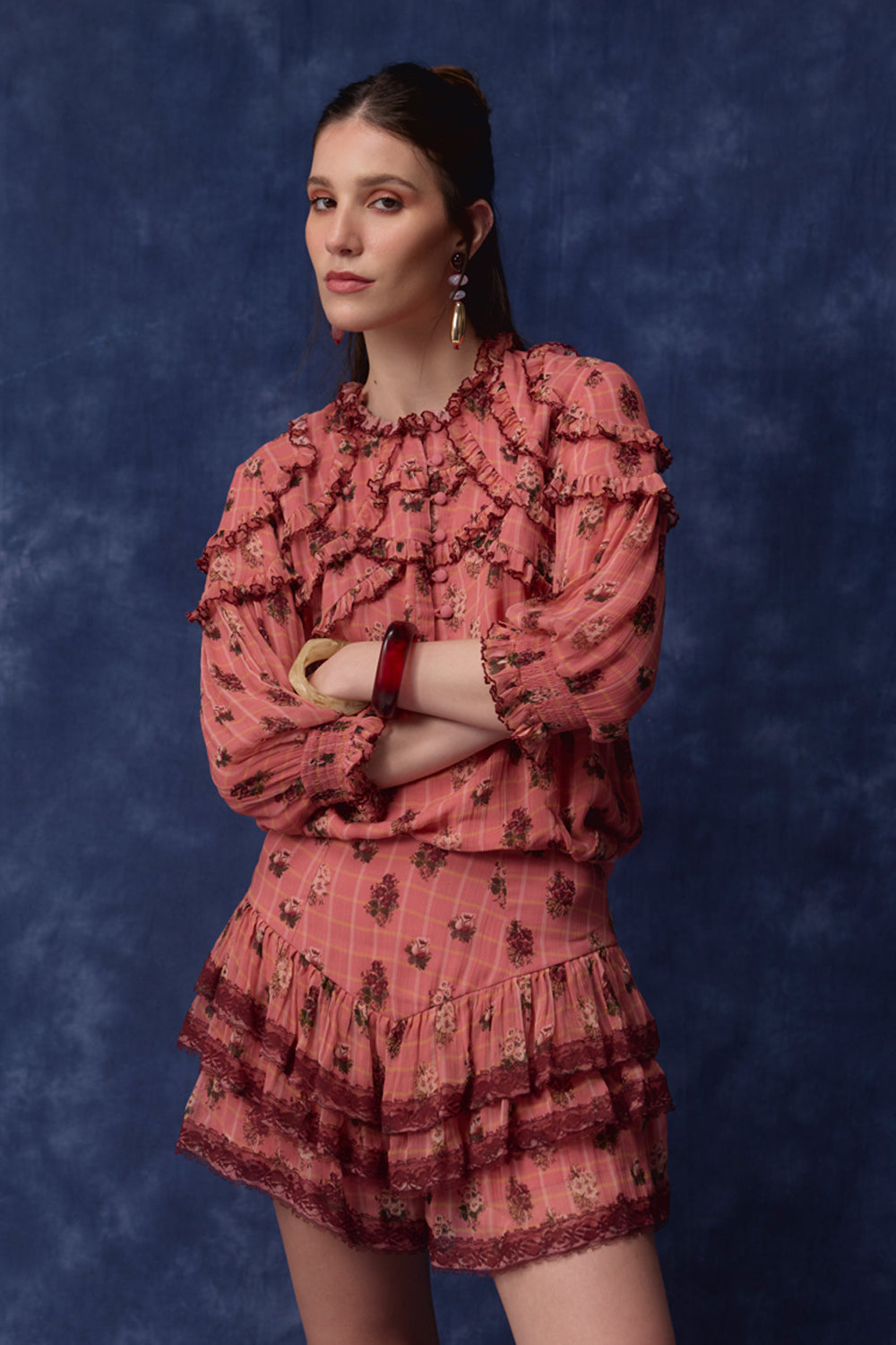 A woman modeling a matching ruffled blouse and shorts set in dusty rose pink with a dark floral pattern, posed against a textured dark blue studio backdrop.
