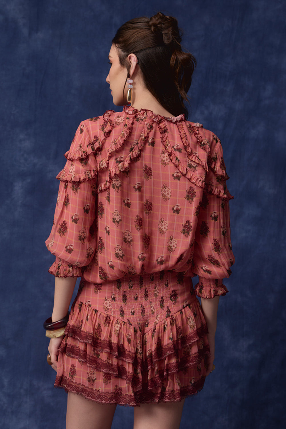 A woman shown from the back wearing a dusty rose pink ruffled blouse and matching mini skirt set with a dark floral print, posed against a textured dark blue studio backdrop.