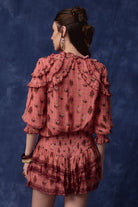 A woman shown from the back wearing a dusty rose pink ruffled blouse and matching mini skirt set with a dark floral print, posed against a textured dark blue studio backdrop.