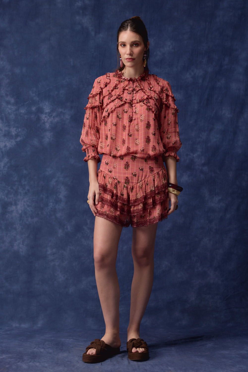 A woman stands against a mottled blue studio backdrop wearing a matching two-piece outfit with a dusty rose and dark floral print, featuring a ruffled long-sleeve top and coordinating shorts, paired with dark brown slide sandals.