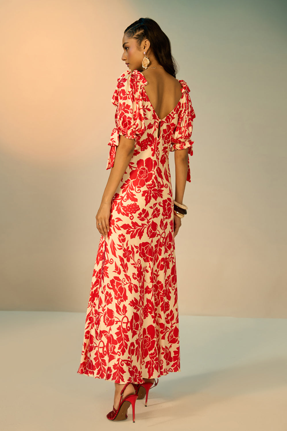 A woman modeling a red and cream floral print maxi dress with a deep V-back and ruffled sleeves, shown from the back against a neutral studio backdrop.