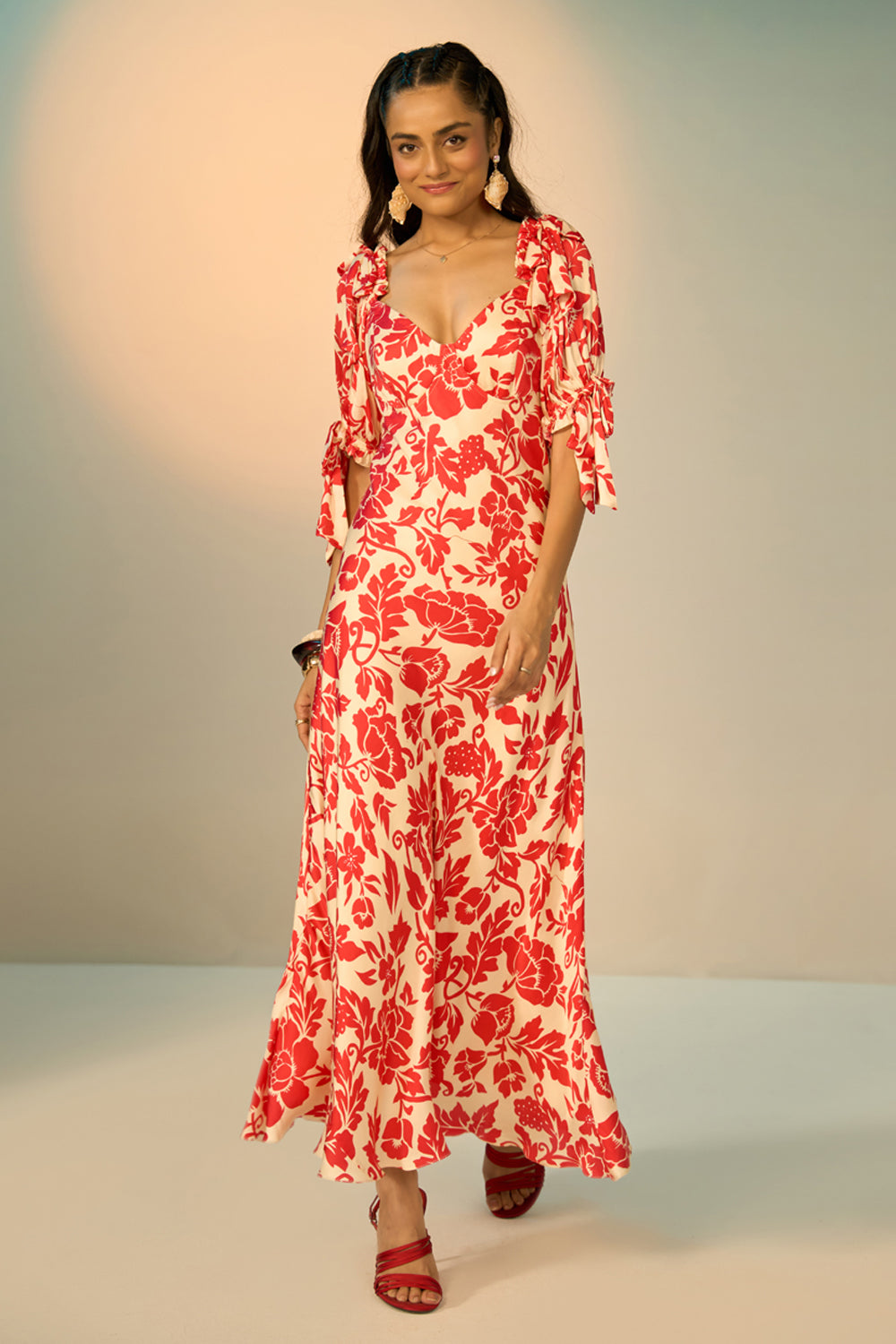 A woman modeling a red and cream floral print maxi dress with ruffled sleeves and a deep V-neckline, standing against a neutral studio backdrop.