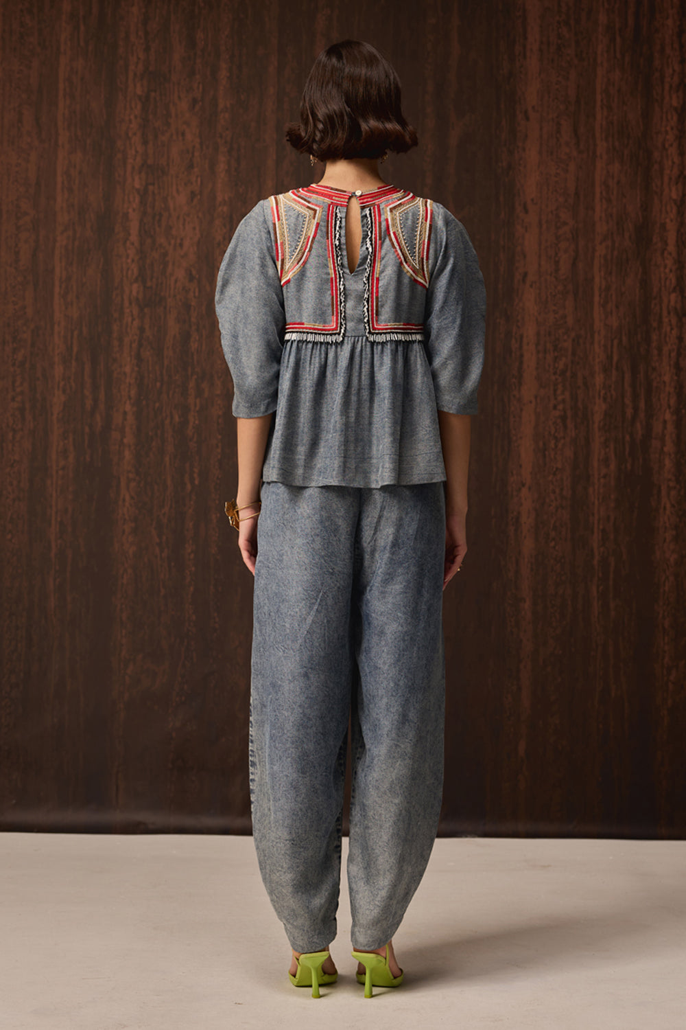 A model viewed from the back wearing a matching gray two-piece set consisting of a short-sleeve peplum top with colorful embroidered detailing on the back and wide-leg pants, set against a dark wood-paneled background.