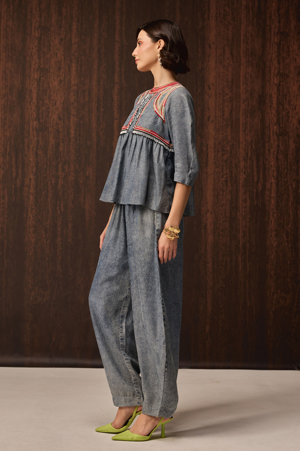A woman stands in profile wearing a light blue denim two-piece outfit with embroidered detailing on the top and wide-leg pants, paired with lime green heels, against a dark wood-paneled background.