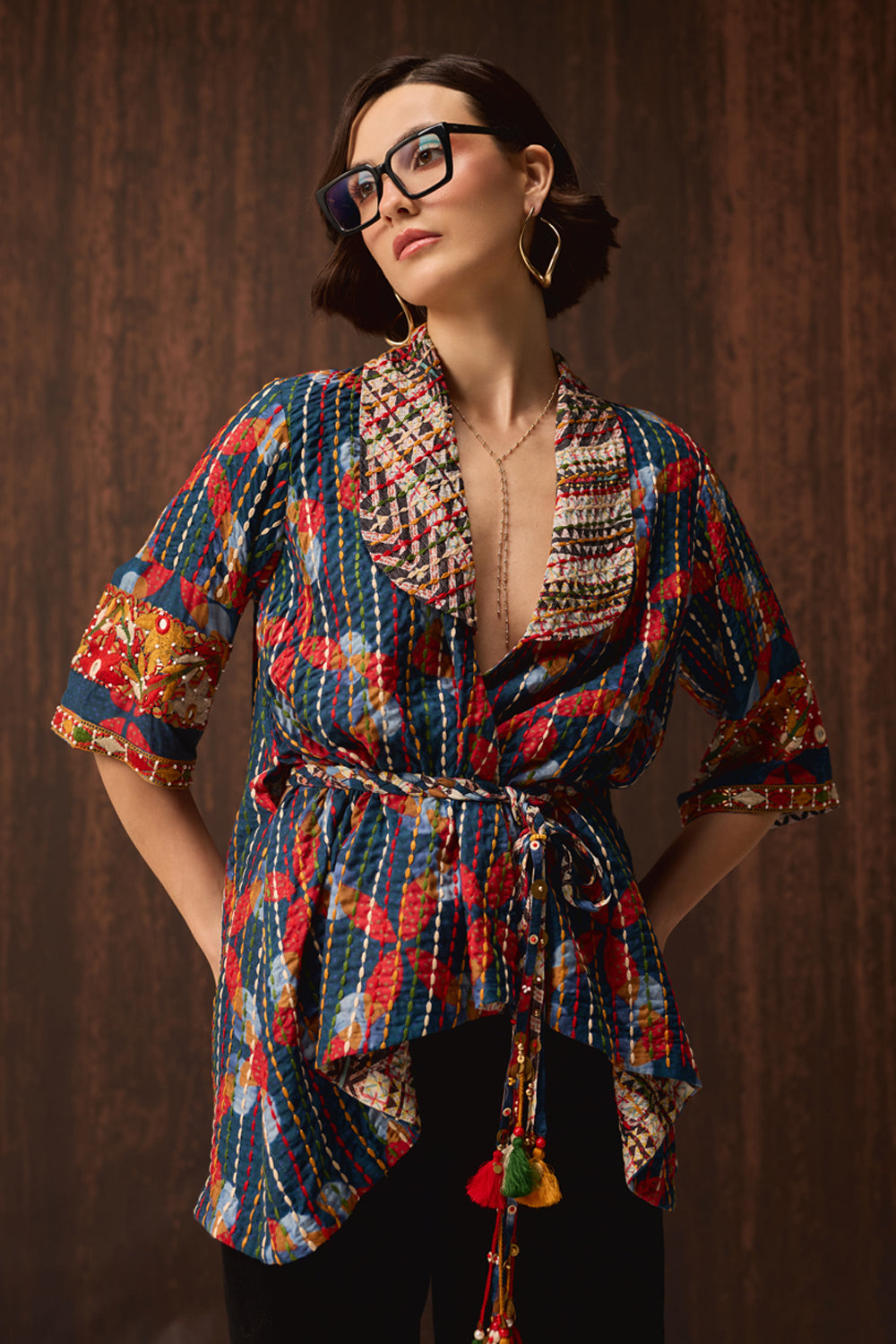 A woman wearing a colorful patterned wrap-style blouse with a tie waist and tassel detail, paired with black pants, posed against a dark wood-paneled background.