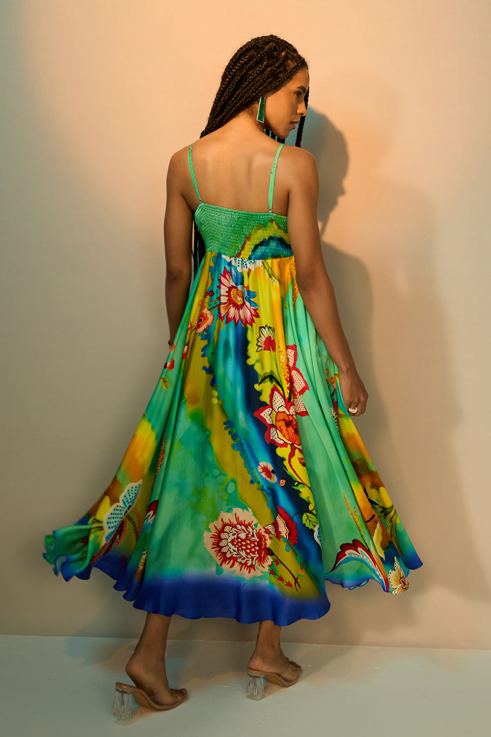 A woman wearing a vibrant, multicolored floral maxi dress, shown from behind as she twirls to display the back of the garment. The dress features a green bodice and a flowing skirt with a colorful print of flowers and tropical motifs in shades of blue, yellow, green, and red.