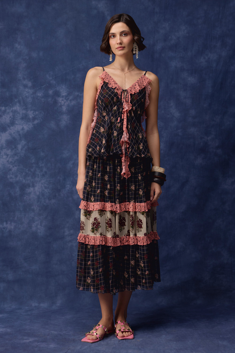 A woman models a bohemian-style tiered maxi dress with a black and navy plaid pattern, pink lace trim, and a central cream panel with floral embroidery. She is standing against a mottled blue studio backdrop.