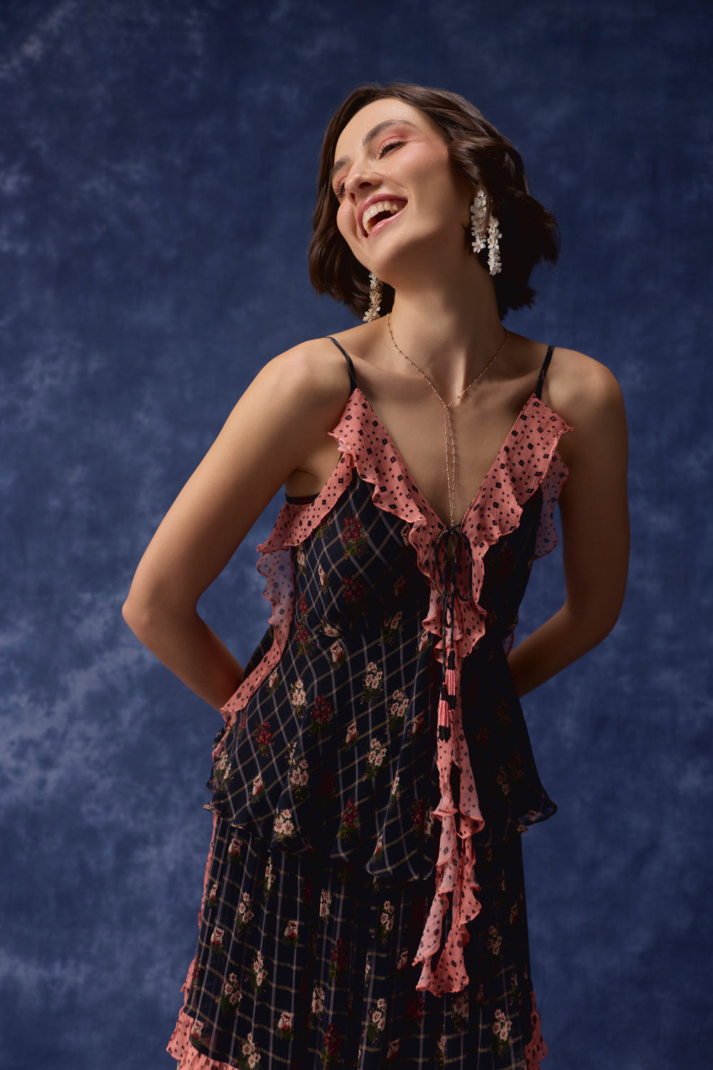 A woman wearing a dark navy and pink floral slip dress with ruffled trim, posing against a textured dark blue backdrop.