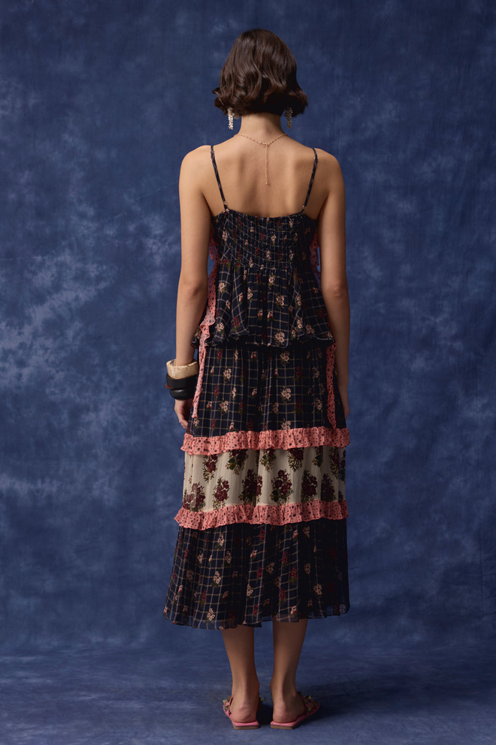A woman is shown from behind wearing a dark navy plaid dress with pink ruffle trim, posing with her arms raised against a textured dark blue backdrop.