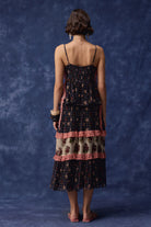 A woman is shown from behind wearing a dark navy plaid dress with pink ruffle trim, posing with her arms raised against a textured dark blue backdrop.