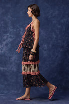 A woman modeling a sleeveless tiered maxi dress with a dark plaid pattern, cream floral inserts, and pink ruffle trim, posed against a textured blue studio backdrop.