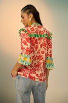 A woman shown from the back wearing a red and cream floral blouse with green and yellow striped ruffle trim and striped cuffs, paired with light blue jeans, against a warm beige studio background.