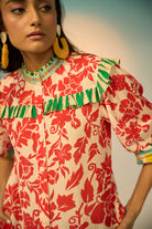 A close-up of a woman wearing a cream-colored blouse with a bold red floral print and green ruffle trim along the collar and shoulders. She is also wearing large yellow hoop earrings.