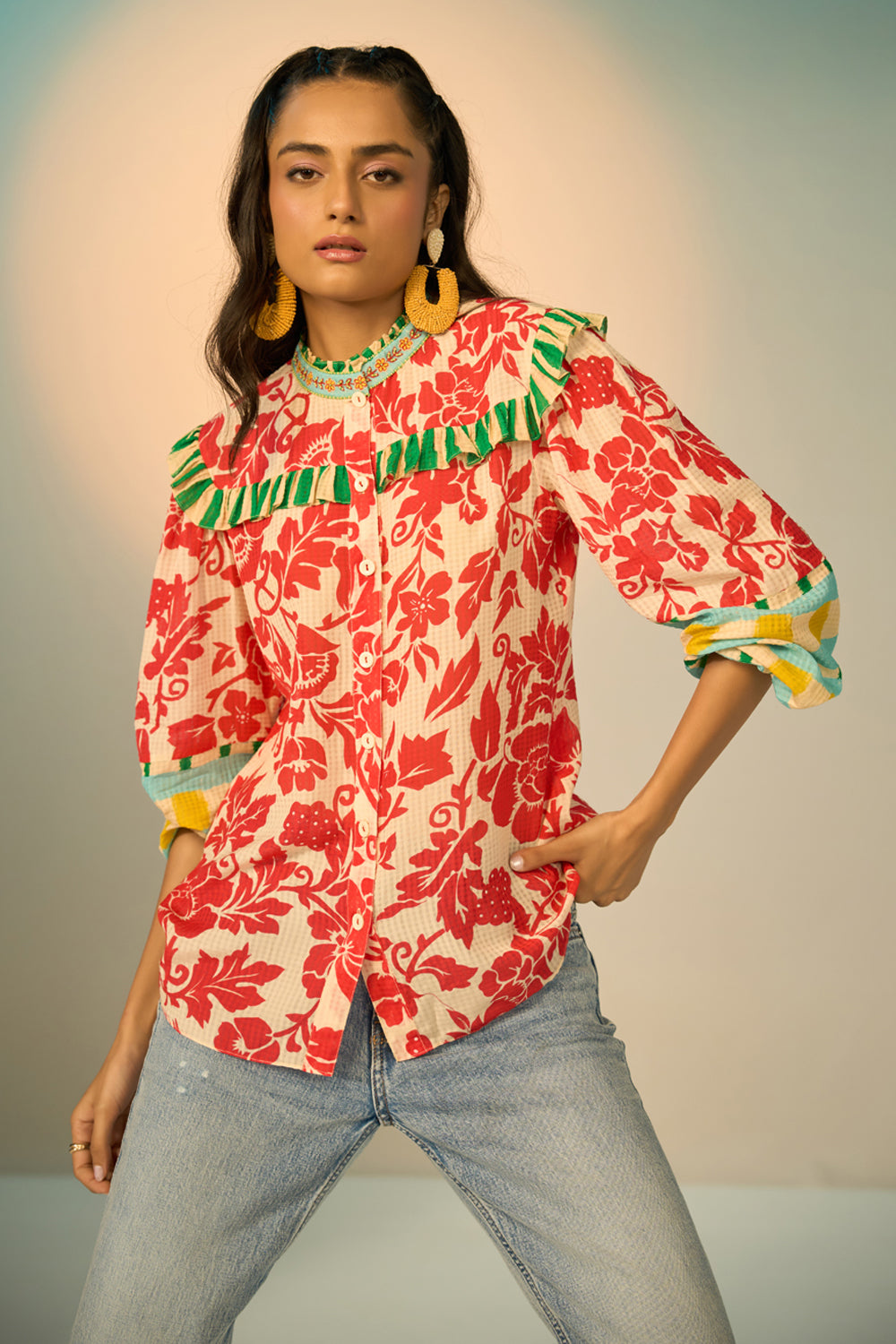 A woman modeling a vibrant red and cream floral print blouse with green and yellow ruffle details, paired with light-wash jeans and gold hoop earrings, set against a plain light gray studio background.