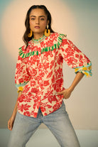 A woman modeling a vibrant red and cream floral print blouse with green and yellow ruffle details, paired with light-wash jeans and gold hoop earrings, set against a plain light gray studio background.