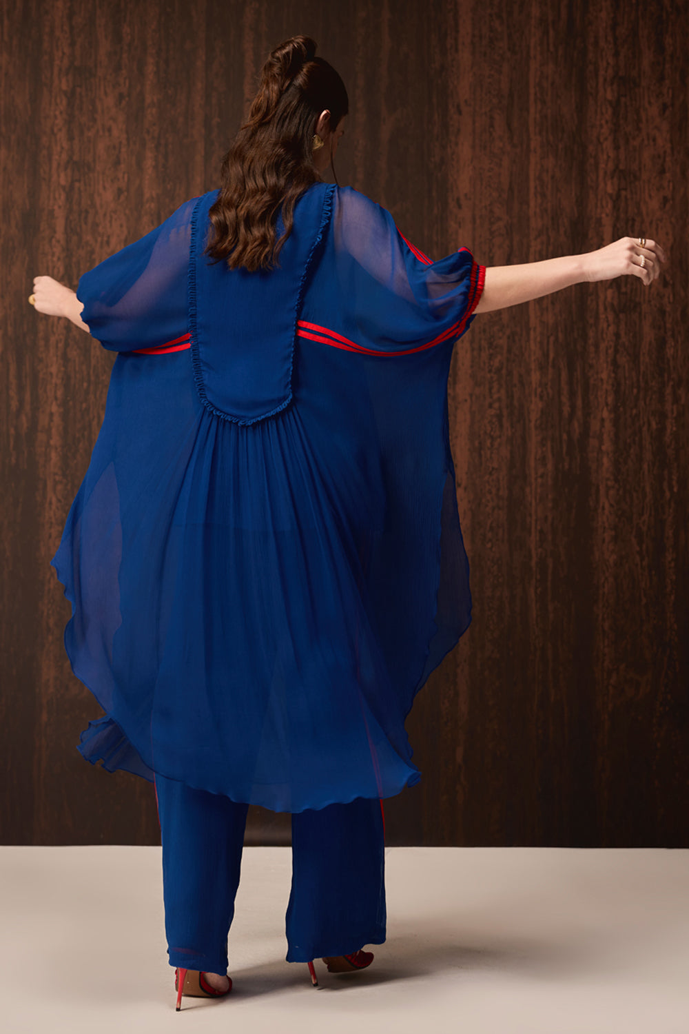 A woman viewed from behind wearing a flowing royal blue sheer tunic with red trim over matching blue pants, posed against a dark wood-paneled background.