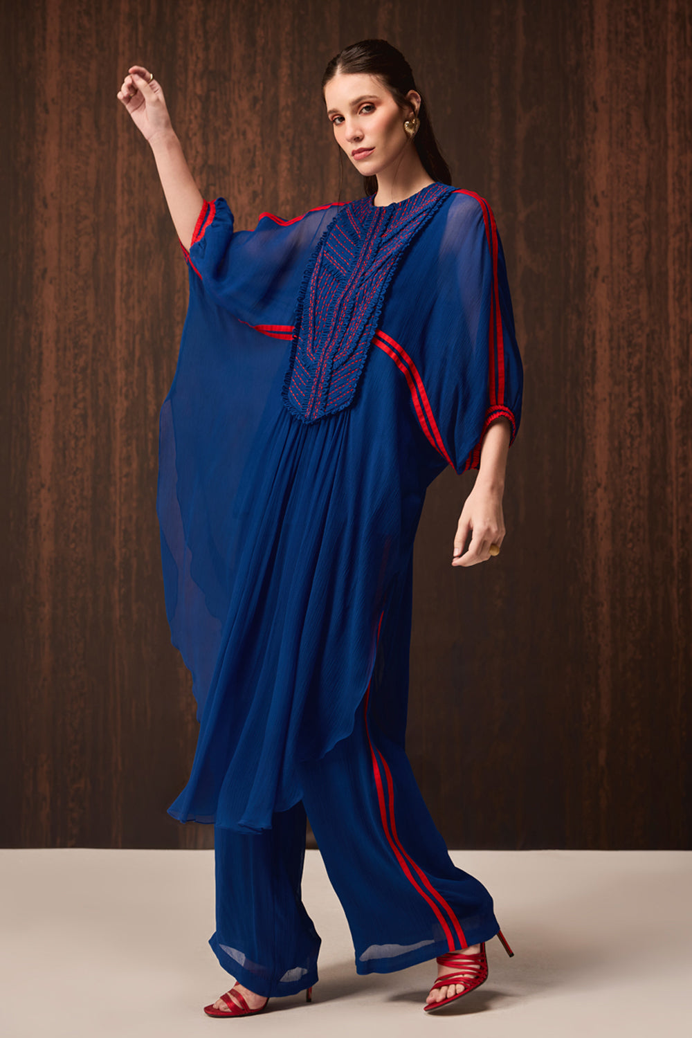 A woman models a royal blue traditional outfit with red stripe accents and a heavily embroidered neckline, set against a dark wood-paneled background.
