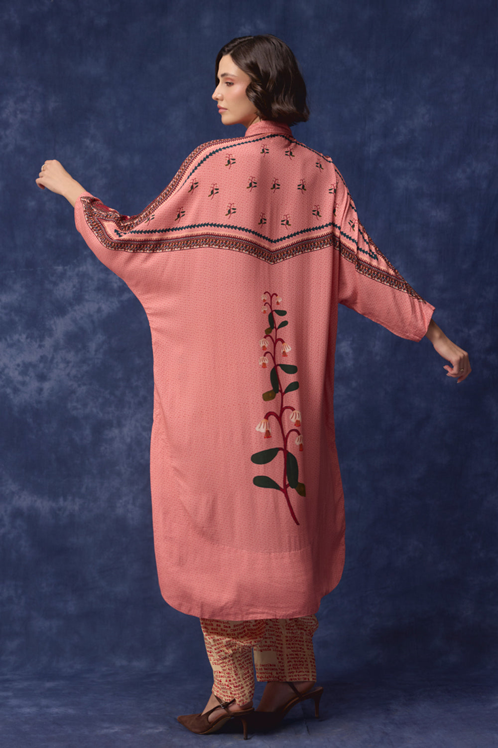 A woman modeling a dusty pink embroidered kurta from the back, featuring a vertical floral vine motif and decorative borders, set against a textured blue studio backdrop.